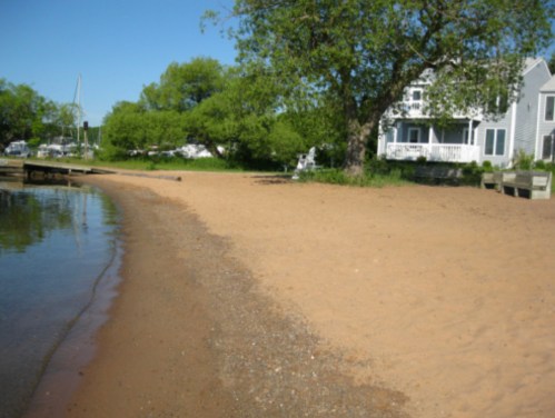 A sandy beach beside a calm waterway, with green trees and a house in the background under a clear blue sky.