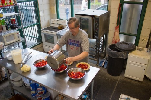 A man prepares food in a kitchen, pouring ingredients into bowls filled with strawberries on a stainless steel table.