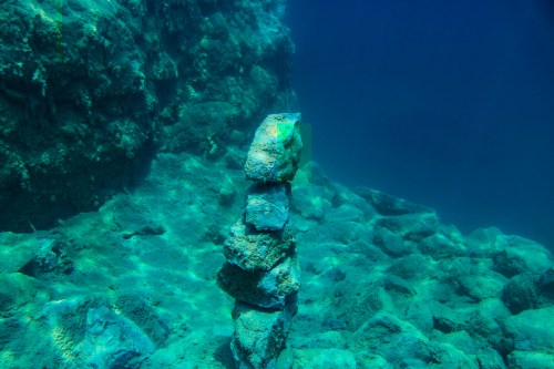 A stack of rocks underwater, surrounded by clear blue water and rocky seabed.