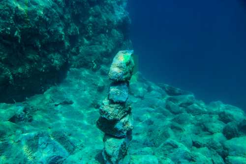 A stack of rocks underwater, surrounded by clear blue water and rocky seabed.