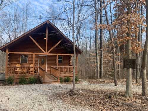 A cozy log cabin surrounded by trees, featuring a porch with chairs and a gravel path leading to a sign.