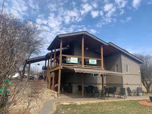 A cozy café with a wooden deck, surrounded by trees and a grassy area, under a partly cloudy sky.