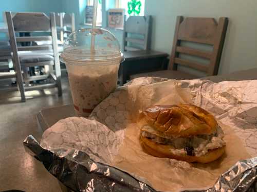 A sandwich wrapped in paper sits on a table next to a frosty drink in a casual dining setting.