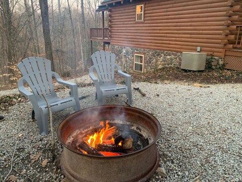 A cozy fire pit with flames, surrounded by two gray chairs, near a log cabin in a wooded area.