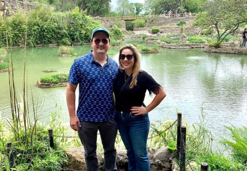 A man and woman stand together by a serene pond surrounded by greenery, smiling at the camera.