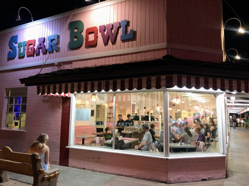 A brightly lit diner named "Sugar Bowl" with people inside, viewed from outside at night.