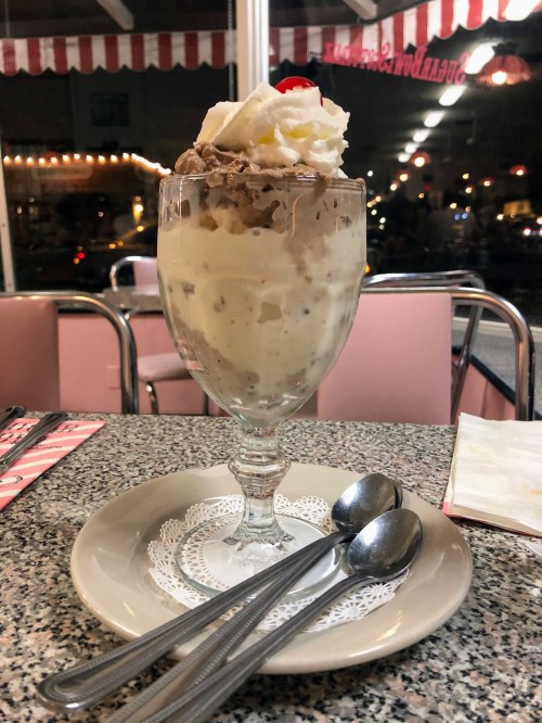 A tall glass dessert with whipped cream and chocolate, served on a plate with two spoons in a retro diner setting.