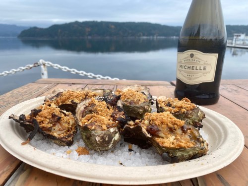 A plate of baked oysters topped with breadcrumbs, served on a bed of salt, with a bottle of sparkling wine in the background.