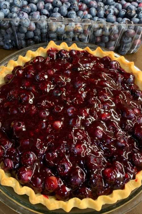 A freshly baked blueberry pie with a glossy berry topping, surrounded by containers of fresh blueberries in the background.