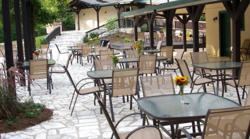Outdoor patio with stone flooring, tables, and chairs, decorated with flowers, surrounded by greenery and buildings.