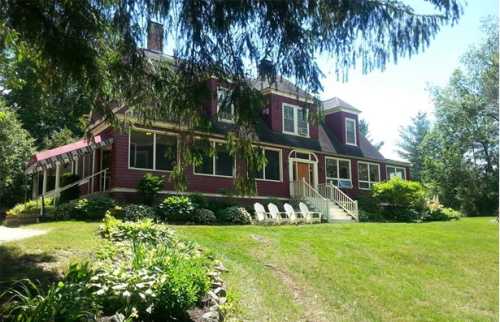 A large, red house with a porch surrounded by greenery and lawn chairs on a sunny day.