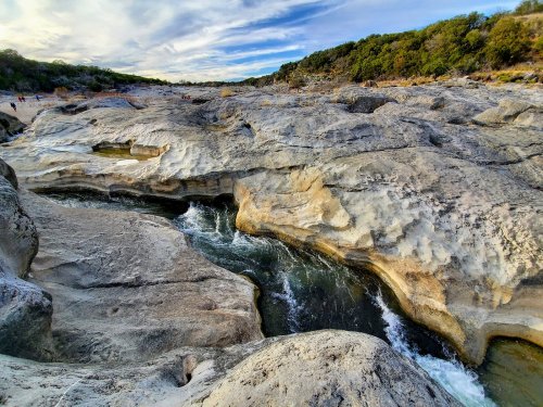 A rocky landscape with a flowing river, surrounded by greenery and a cloudy sky.