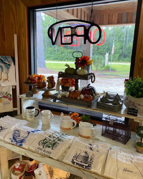 A cozy shop display with seasonal decor, mugs, and table linens, featuring a neon "Open" sign in the background.