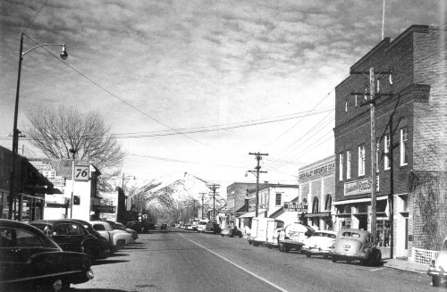 Black and white photo of a small town street lined with vintage cars, shops, and mountains in the background.