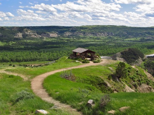 A scenic view of a winding path leading to a rustic cabin on a hill, surrounded by lush greenery and rolling hills.