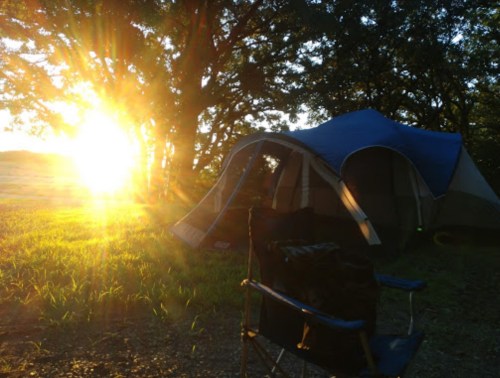 A tent and camping chair are set up in a grassy area, with the sun rising through trees in the background.
