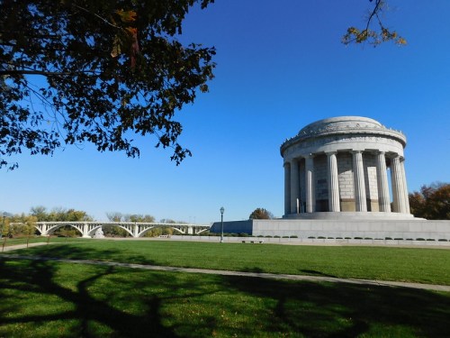 A large stone monument with a domed roof, surrounded by green grass and trees, under a clear blue sky.
