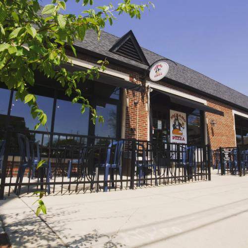 Exterior view of a pizza restaurant with a brick facade, large windows, and outdoor seating under a clear blue sky.