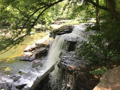 A serene waterfall cascading over rocky ledges, surrounded by lush green trees and a calm river below.