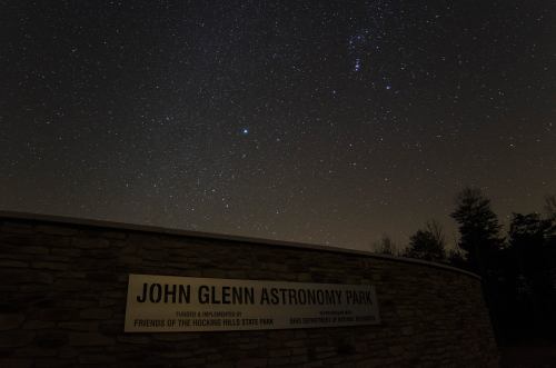 A night sky filled with stars above the John Glenn Astronomy Park sign, surrounded by trees.