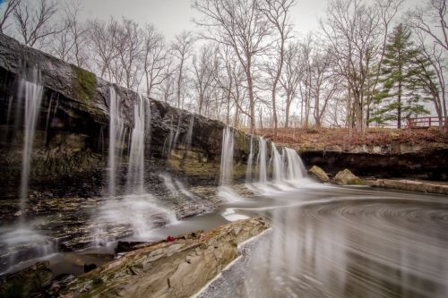 A serene waterfall cascades over rocky ledges, surrounded by bare trees and a calm, reflective pool.