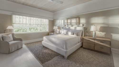 Bright, serene bedroom with a large window, white bed, wooden nightstands, and soft decor.