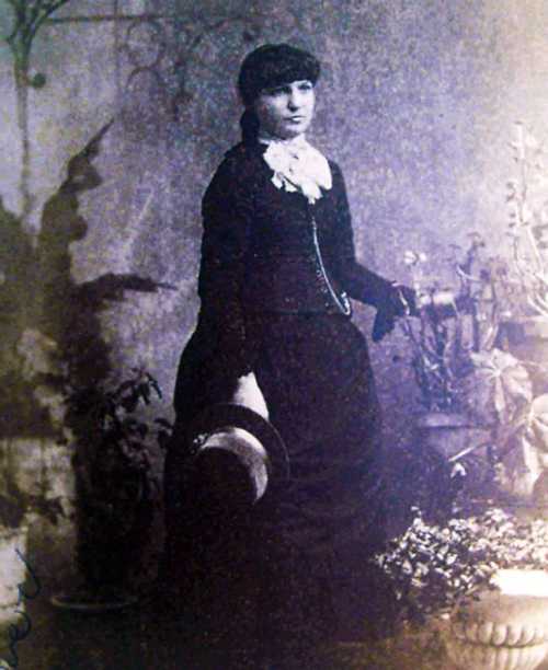 A vintage black-and-white portrait of a woman in a formal dress, holding a hat, surrounded by plants.