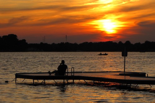 A person sits on a dock at sunset, with vibrant orange and purple skies reflecting on the water.