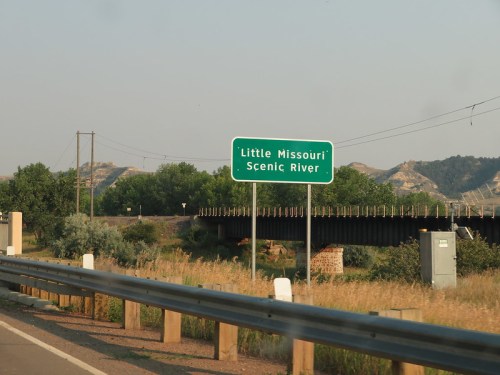 Sign for Little Missouri Scenic River along a highway, with hills and greenery in the background.
