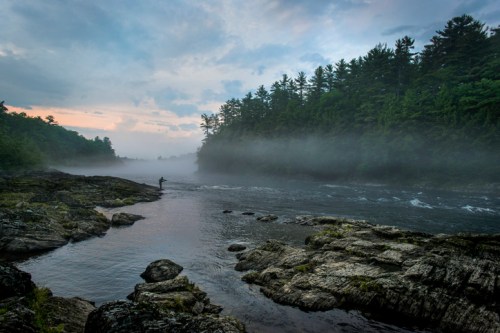 A misty river scene with a lone figure fishing on rocky banks, surrounded by lush trees and a cloudy sky.