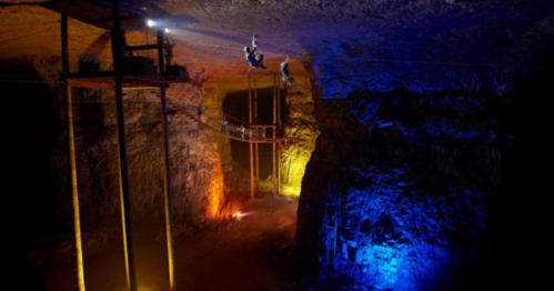 Two people ziplining through a brightly lit underground cavern with colorful lights illuminating the rock formations.