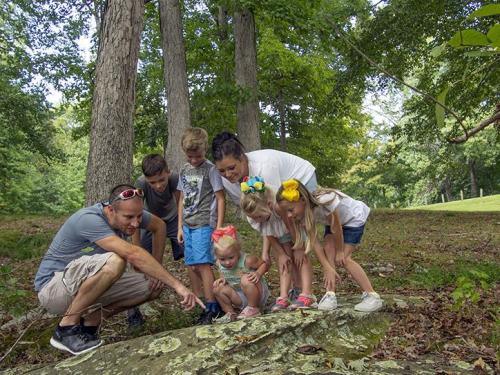 A group of children and adults gather around a rock in a forest, examining something on the ground with curiosity.