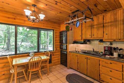 Cozy wooden kitchen with a dining table, large windows, and rustic cabinets, surrounded by greenery outside.