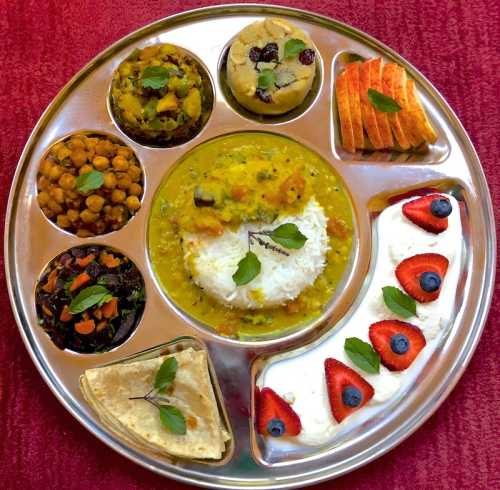 A colorful thali featuring rice, dal, vegetables, chapati, dessert, and fresh fruit, garnished with mint leaves.