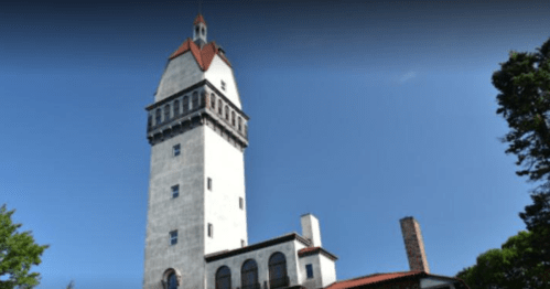 A tall, historic stone tower with a red roof, surrounded by trees and a clear blue sky.