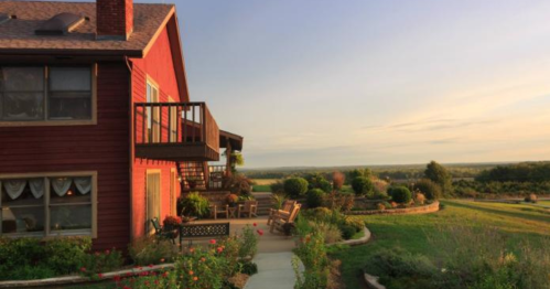 A red house with a balcony overlooks a scenic landscape at sunset, surrounded by gardens and greenery.