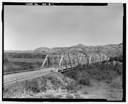 A metal arch bridge spans a river, surrounded by rolling hills and vegetation under a clear sky.