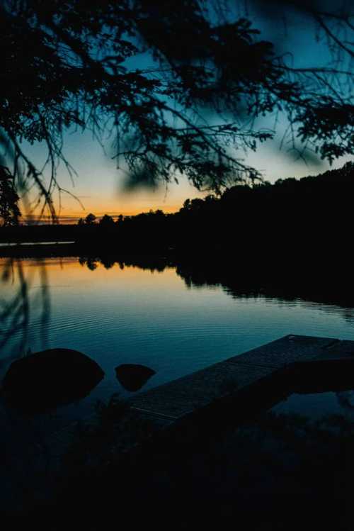 A serene lake at sunset, with silhouettes of trees and a wooden dock reflecting on the calm water.