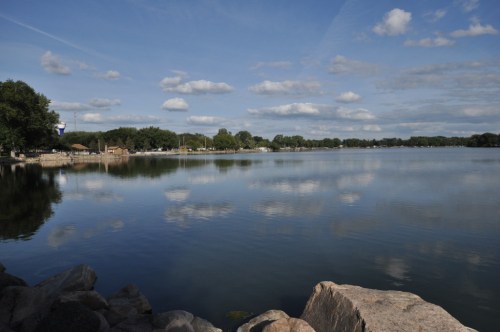 A serene lake scene with calm water reflecting clouds and trees, framed by rocks along the shore.