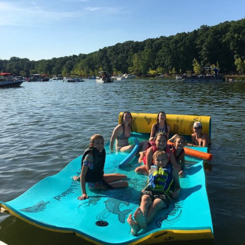A group of children and teens enjoying a floating mat on a lake, surrounded by boats and greenery.