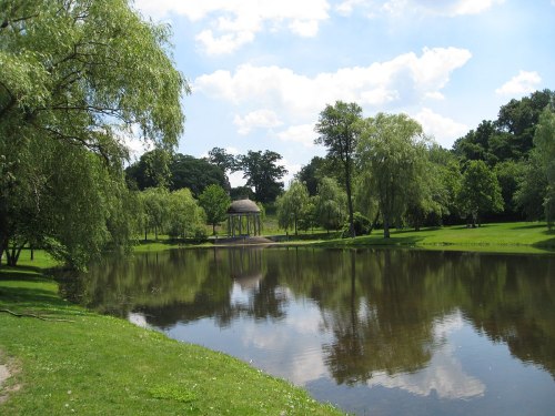 A serene park scene featuring a pond, lush greenery, and a gazebo reflected in the water under a blue sky.