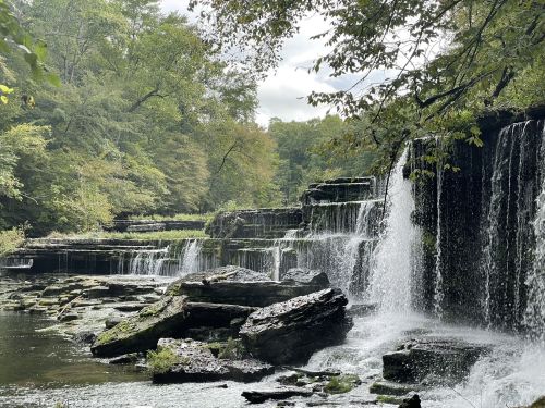 A serene waterfall cascades over rocky ledges, surrounded by lush green trees and a tranquil river.