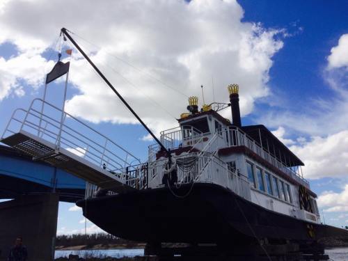 A large, white riverboat with a black and yellow design, docked under a blue sky with fluffy clouds.