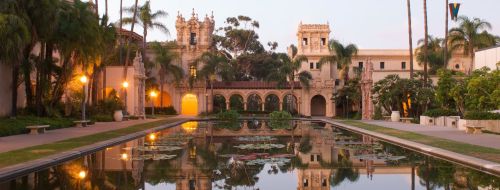A serene view of a landscaped garden with palm trees and a reflecting pond, featuring historic buildings in the background.