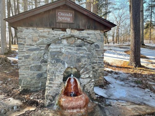 A stone spring structure with a wooden roof, labeled "Orenda Spring," surrounded by trees and remnants of snow.