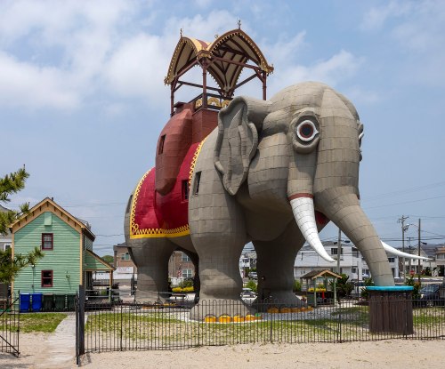 A large, whimsical elephant-shaped building with a tower, surrounded by a fence and nearby colorful houses.