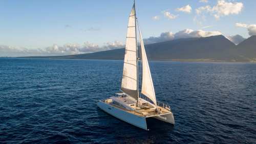 A sailboat with white sails glides over calm blue waters, with mountains and clouds in the background.