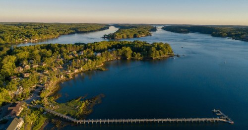 Aerial view of a serene river surrounded by lush greenery and a wooden pier extending into the water.
