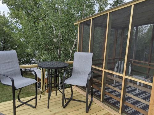 A wooden deck with two gray chairs and a small table, surrounded by trees and a screened-in area.