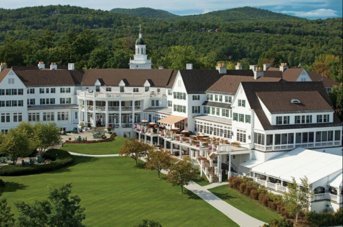 A large, elegant hotel surrounded by green trees and mountains, featuring a spacious lawn and outdoor seating areas.